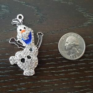 May include: A silver charm featuring a cartoon snowman with a blue scarf and a white, sparkly body. The charm is about the size of a US quarter.
