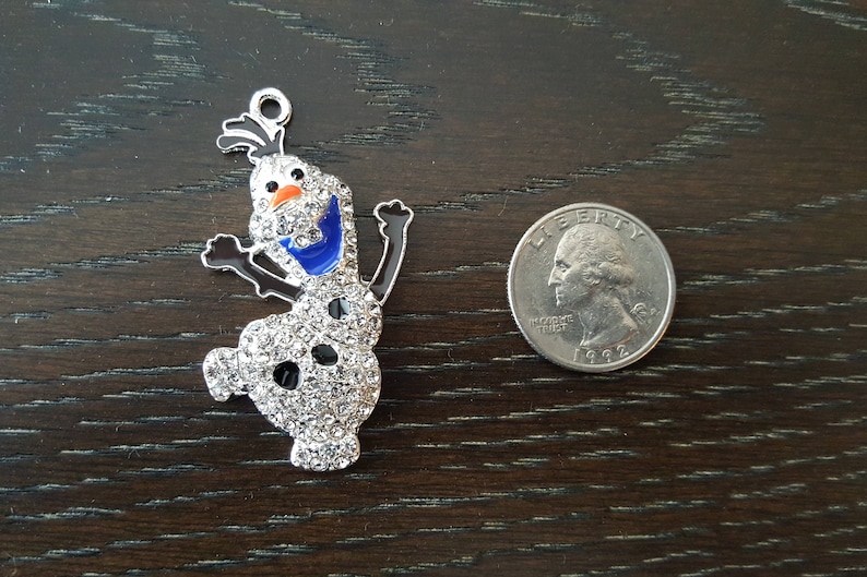 May include: A silver charm featuring a cartoon snowman with a blue scarf and a white, sparkly body. The charm is about the size of a US quarter.