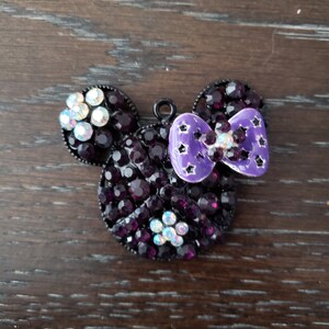 Pu&ograve; includere: Un ciondolo Mickey Mouse in strass nero e viola con un fiocco viola. Il fiocco ha stelle bianche.