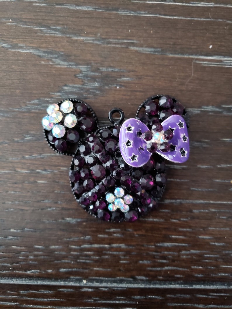 Pu&ograve; includere: Un ciondolo Mickey Mouse in strass nero e viola con un fiocco viola. Il fiocco ha stelle bianche.