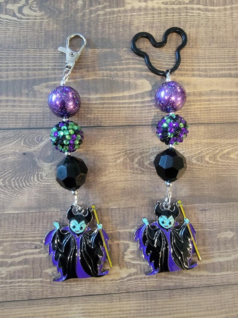 May include: Two beaded keychains. One has a silver clasp, the other a black Mickey Mouse shape. Each keychain features purple, green, and black beads, and a charm of a cartoon villain in black and purple robes, holding a yellow staff.