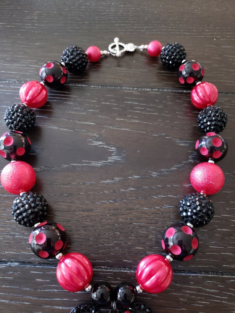 May include: A black and red chunky necklace with a large black and red ladybug charm. The necklace is made of large, round beads with a variety of textures, including smooth, textured, and sparkly.