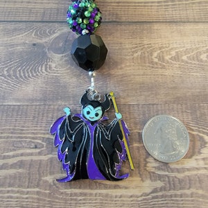 May include: A beaded keychain featuring a purple and black Maleficent charm. The charm is a detailed enamel design with black, purple, and yellow accents. The keychain includes a series of decorative beads in purple, green, and black.