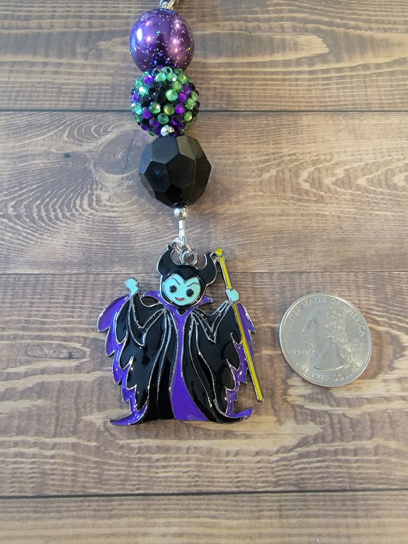 May include: A beaded keychain featuring a purple and black Maleficent charm. The charm is a detailed enamel design with black, purple, and yellow accents. The keychain includes a series of decorative beads in purple, green, and black.