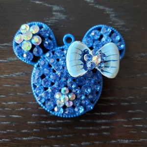 May include: A blue rhinestone charm shaped like Mickey Mouse with a light blue bow. The charm has a small loop at the top for attaching to a necklace or bracelet.