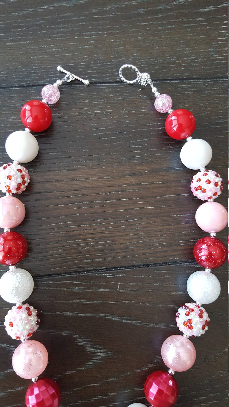 Pode incluir: Um colar de contas grossas vermelho, branco e rosa com fechos prateados. As contas s&atilde;o grandes e t&ecirc;m uma variedade de texturas, incluindo lisas, brilhantes e texturizadas.