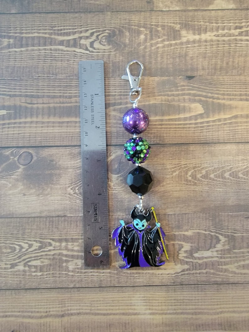 May include: A beaded keychain featuring a cartoon image of Maleficent. The keychain has a silver clasp, purple and green beads, and a black geometric bead. The Maleficent charm is black and purple.