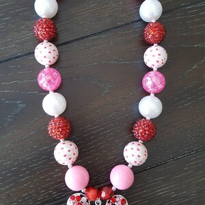 May include: A pink and red beaded necklace with a Minnie Mouse charm. The beads are a mix of solid colors and white beads with red hearts.
