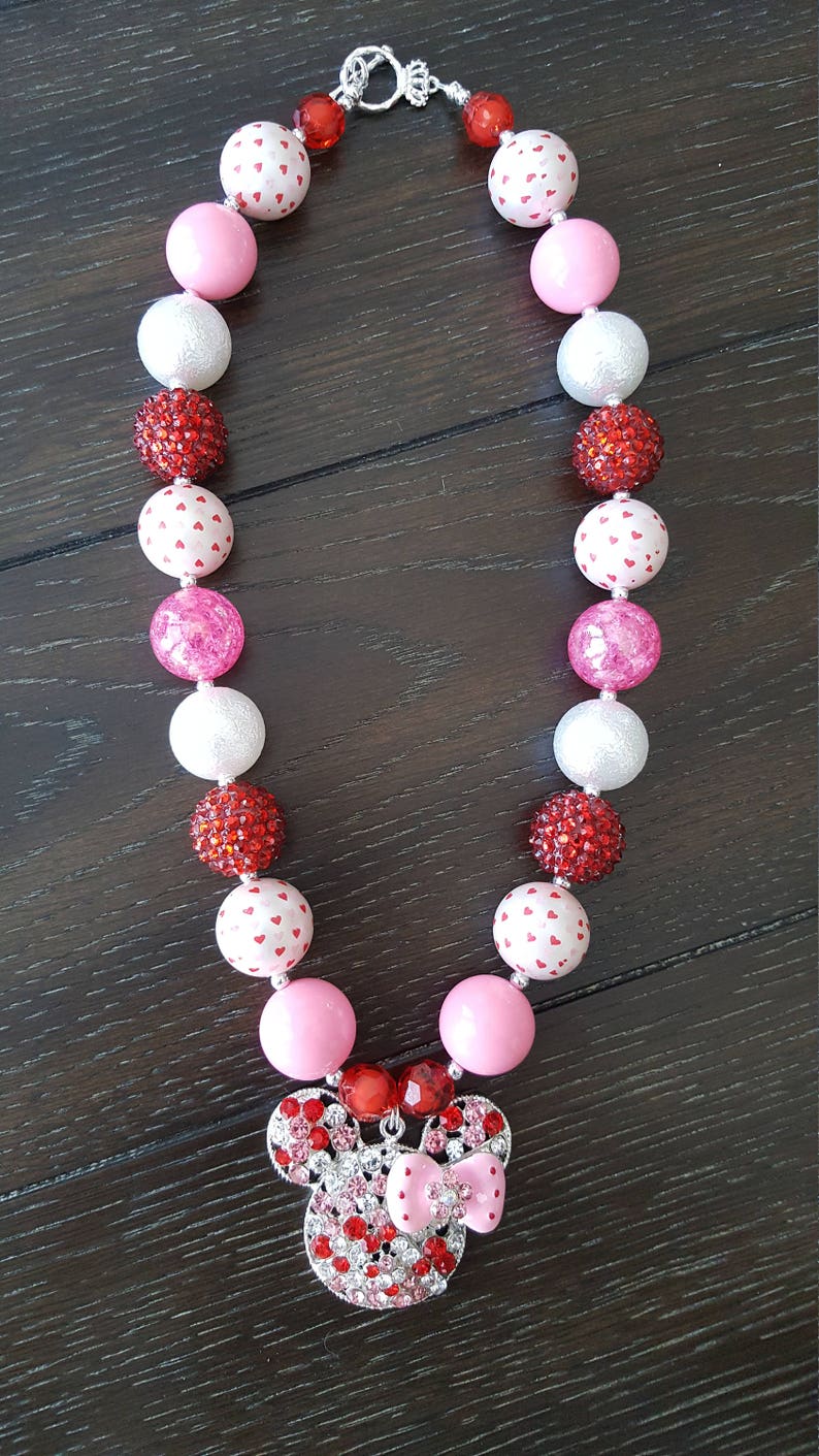 May include: A pink and red beaded necklace with a Minnie Mouse charm. The beads are a mix of solid colors and white beads with red hearts.