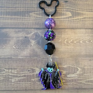 May include: A black Mickey Mouse-shaped keychain with a series of beads in purple, black, and green, culminating in a Maleficent charm. The charm features the character in black and purple with a yellow staff.