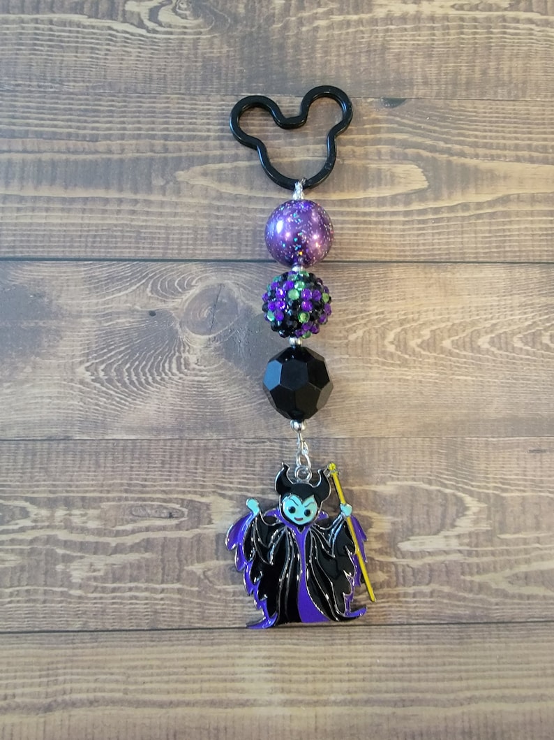 May include: A black Mickey Mouse-shaped keychain with a series of beads in purple, black, and green, culminating in a Maleficent charm. The charm features the character in black and purple with a yellow staff.