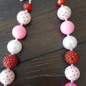 May include: A Valentine's Day themed chunky necklace with alternating pink, white, and red beads. The beads are decorated with white hearts and rhinestones. The necklace has a silver toggle clasp.