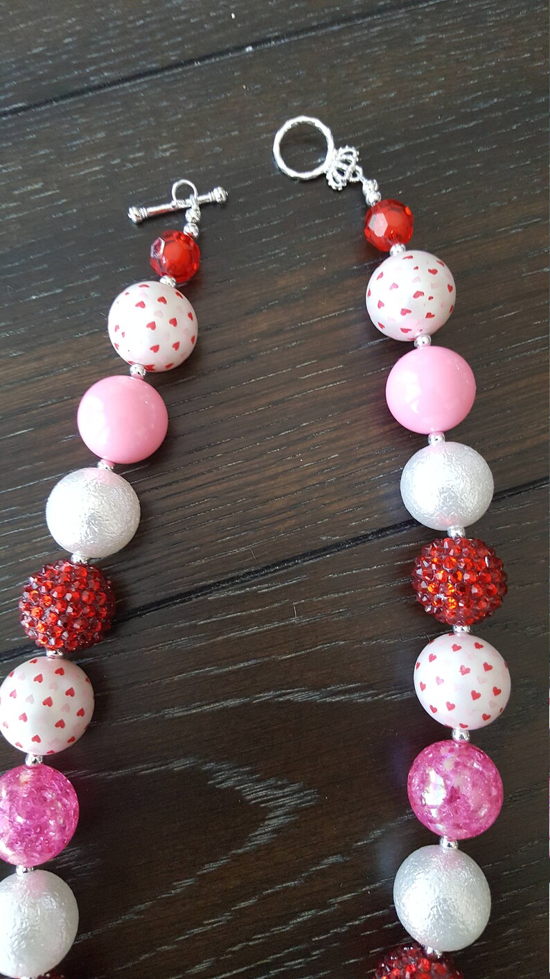 May include: A Valentine's Day themed chunky necklace with alternating pink, white, and red beads. The beads are decorated with white hearts and rhinestones. The necklace has a silver toggle clasp.