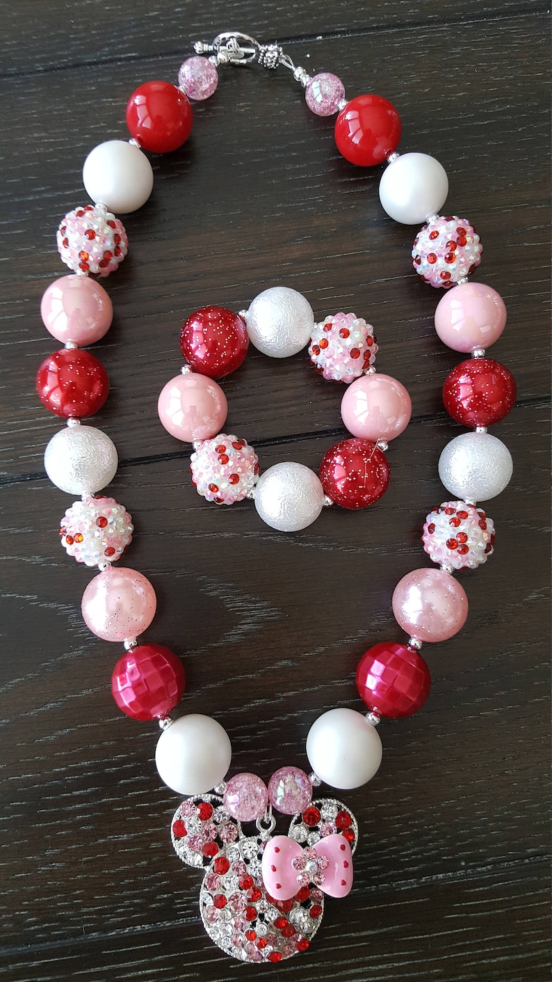Pode incluir: Um colar de contas grossas vermelho, branco e rosa com um fecho prateado e um pingente de Minnie Mouse de strass. O colar vem com uma pulseira combinando.