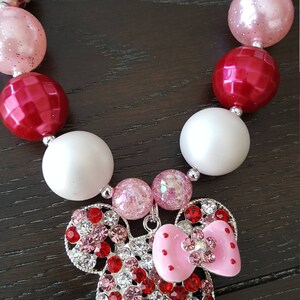 Pode incluir: Um colar de contas vermelhas, brancas e rosadas com um amuleto de Minnie Mouse de strass. O amuleto tem um la&ccedil;o rosa e strass vermelhos e brancos.