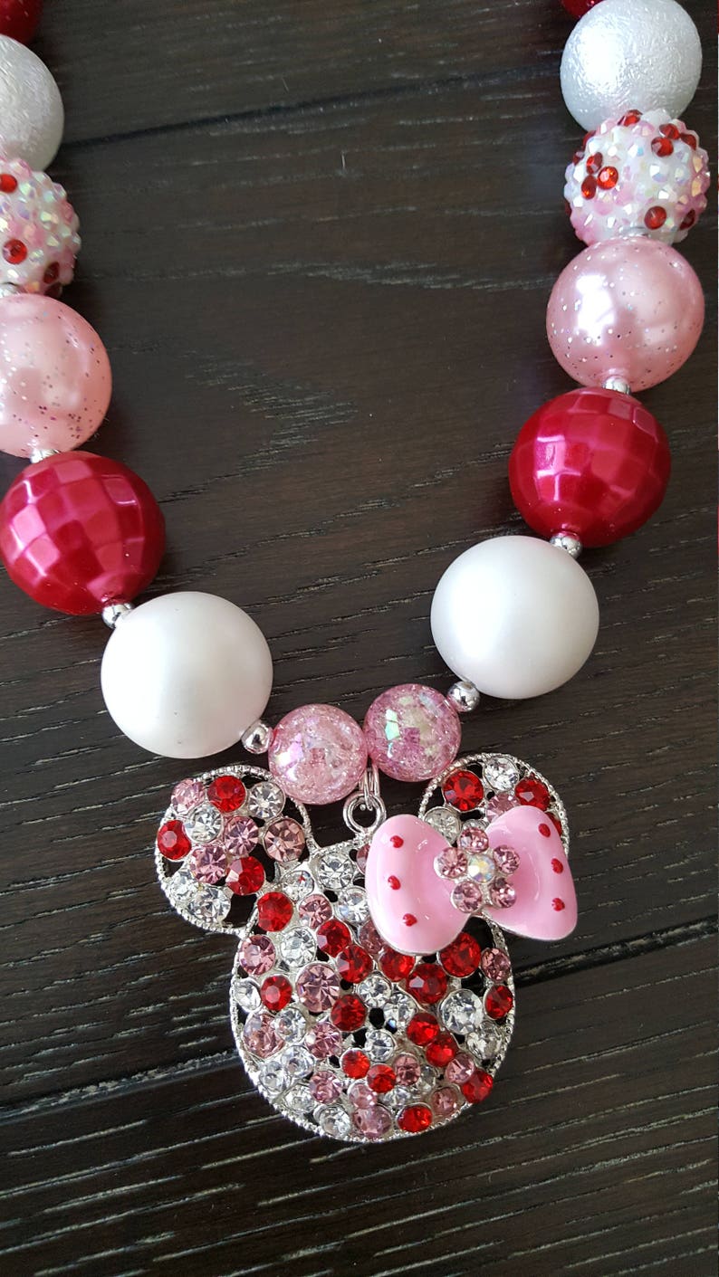Pode incluir: Um colar de contas vermelhas, brancas e rosadas com um amuleto de Minnie Mouse de strass. O amuleto tem um la&ccedil;o rosa e strass vermelhos e brancos.