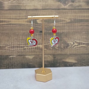 May include: A pair of silver heart-shaped earrings with a red, white, and yellow Kansas City Chiefs logo. The earrings are hanging from a gold stand and have red and yellow beads.