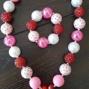 May include: A pink and red beaded necklace and bracelet set with a Minnie Mouse charm. The beads are a mix of solid colors, white with red hearts, and red glitter.