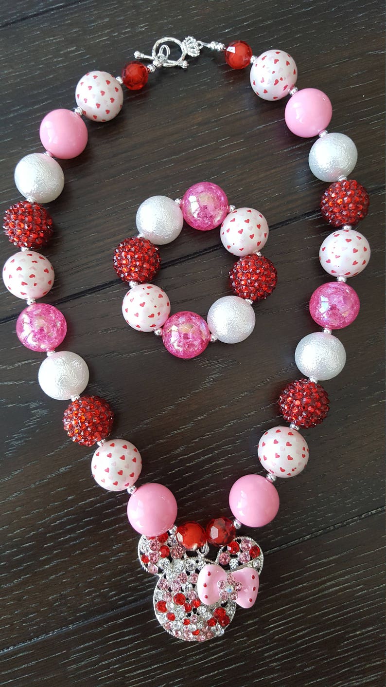 May include: A pink and red beaded necklace and bracelet set with a Minnie Mouse charm. The beads are a mix of solid colors, white with red hearts, and red glitter.