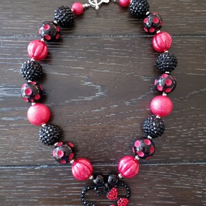 May include: A red and black chunky necklace with a Minnie Mouse charm. The necklace is made of large, round beads with black polka dots and black glitter beads. The Minnie Mouse charm is made of black metal with red rhinestones.