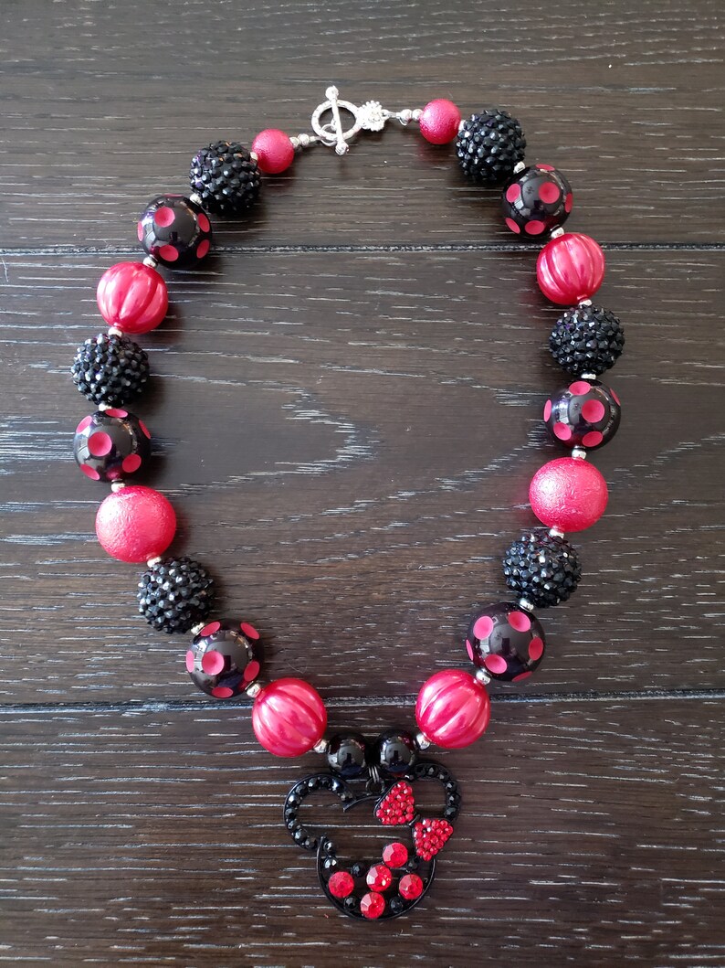 May include: A red and black chunky necklace with a Minnie Mouse charm. The necklace is made of large, round beads with black polka dots and black glitter beads. The Minnie Mouse charm is made of black metal with red rhinestones.