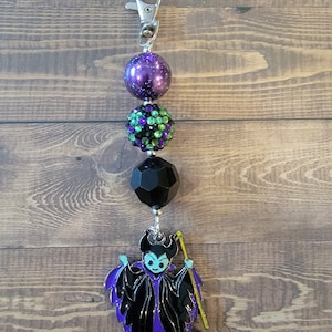 May include: A keychain featuring a silver clasp, purple and green beaded accents, and a black geometric bead. The charm is a stylized image of a character in a black and purple outfit with a yellow staff.