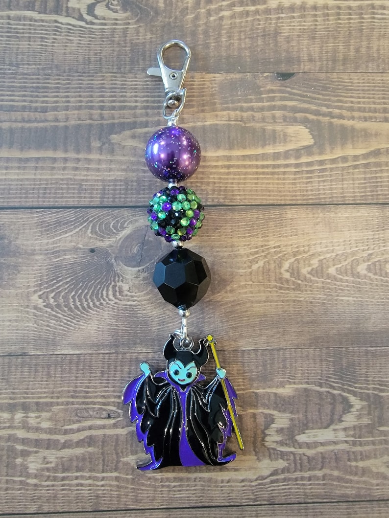 May include: A keychain featuring a silver clasp, purple and green beaded accents, and a black geometric bead. The charm is a stylized image of a character in a black and purple outfit with a yellow staff.
