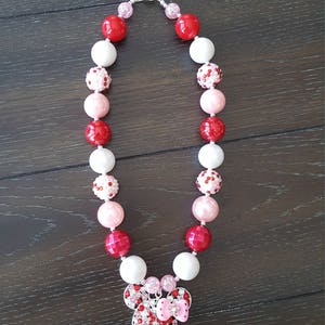 Pode incluir: Um colar de contas grossas vermelho, rosa e branco com um pingente de Minnie Mouse de strass.