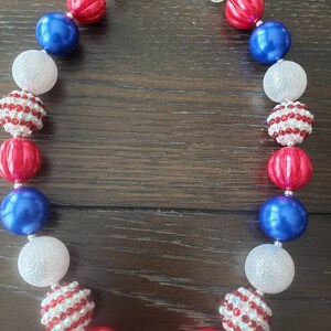 May include: A necklace with red, white, and blue beads. The necklace features a mix of round and textured beads, including shiny red, solid blue, and sparkling white beads. The necklace is secured with a silver-coloured clasp.