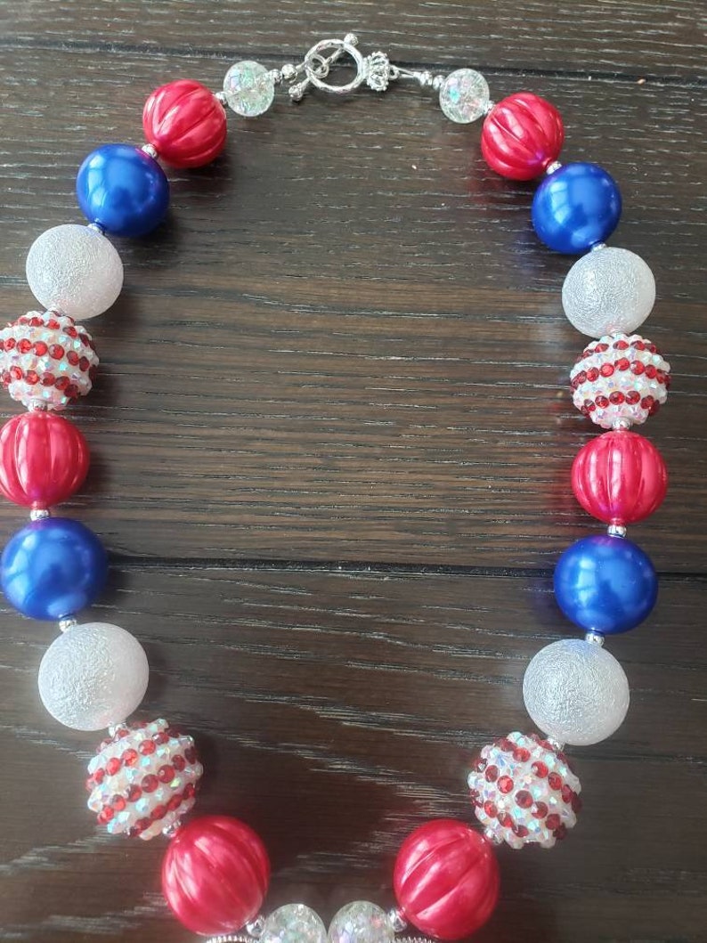 May include: A necklace with red, white, and blue beads. The necklace features a mix of round and textured beads, including shiny red, solid blue, and sparkling white beads. The necklace is secured with a silver-coloured clasp.