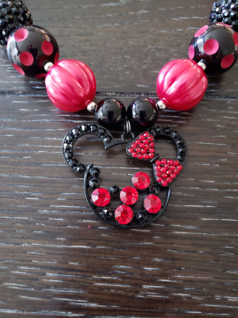 May include: A black and red beaded necklace with a Minnie Mouse charm. The charm is made of black metal and has red rhinestones.