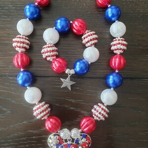 May include: A red, white, and blue beaded necklace and bracelet set. The necklace and bracelet feature large, round beads in red, white, and blue. The necklace has a Mickey Mouse pendant. The bracelet has a star charm that says "dream".