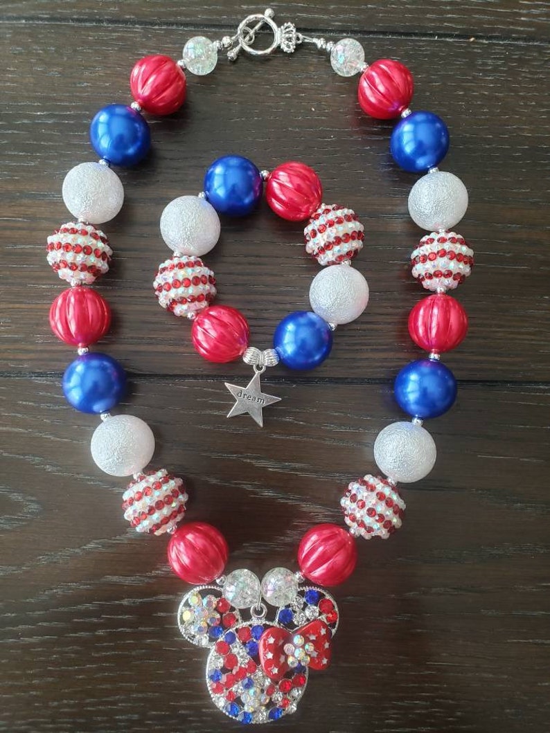 May include: A red, white, and blue beaded necklace and bracelet set. The necklace and bracelet feature large, round beads in red, white, and blue. The necklace has a Mickey Mouse pendant. The bracelet has a star charm that says "dream".