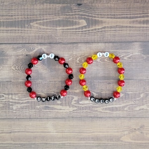 May include: Two beaded bracelets with black letter beads spelling out the names "SWIFT" and "KELCE". The "SWIFT" bracelet has red and black beads, while the "KELCE" bracelet has yellow and red beads.