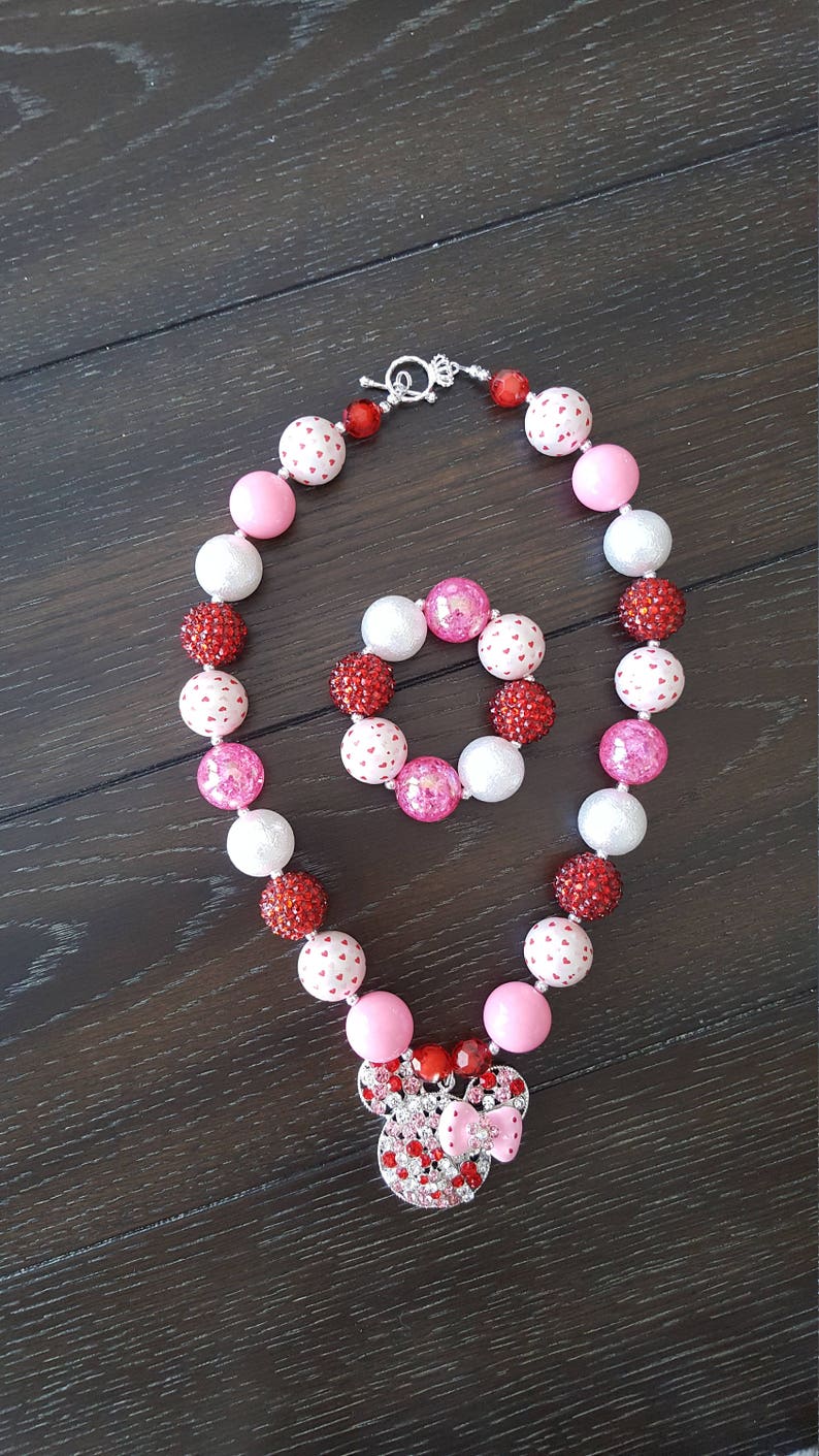 May include: A pink and red beaded necklace and bracelet set with a Minnie Mouse charm. The beads are white with red hearts, pink, and red.