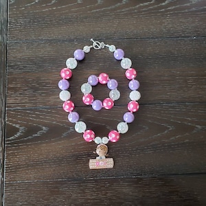 May include: A pink and purple beaded necklace with white polka dots and a silver clasp. The necklace has a charm of a girl with a stethoscope and the word "DOC" on it.