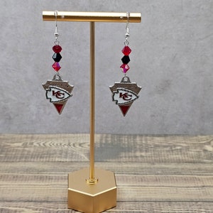 May include: A pair of silver earrings with red and black crystal beads and a Kansas City Chiefs logo charm. The earrings are displayed on a gold stand.