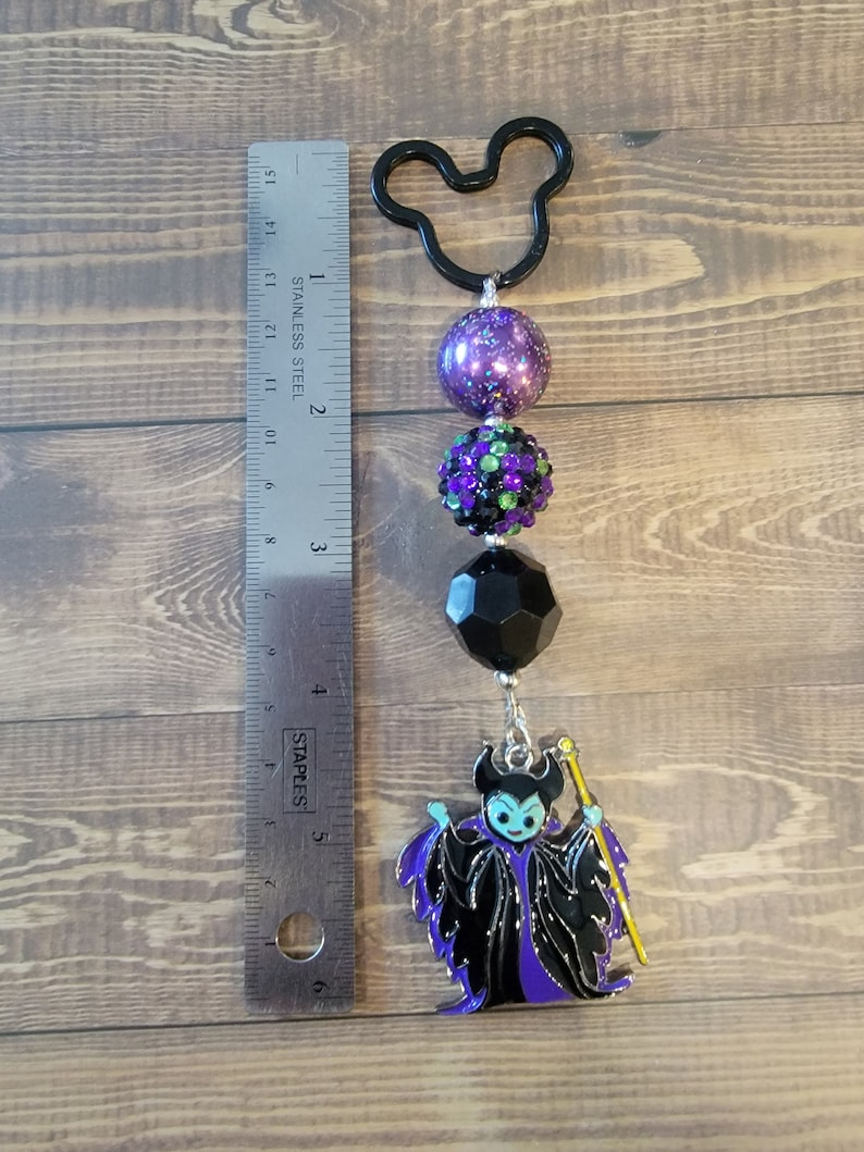 May include: A black Mickey Mouse-shaped keychain with a beaded design. The keychain features a purple glitter bead, a green and black rhinestone bead, a black faceted bead, and a Maleficent charm. The charm is approximately 2 inches in length.