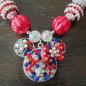 May include: A patriotic necklace with a Mickey Mouse pendant, decorated with red, white, and blue rhinestones. The necklace has large red and white beads, and a red bow with white stars. The pendant is silver-toned.