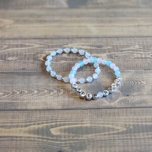 May include: Two beaded bracelets with the words "PAPER RINGS" spelled out in silver beads. The bracelets are made with light blue and pink beads.