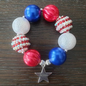 May include: A bracelet with red, white, and blue beads, some plain and some decorated with rhinestones. A silver star charm with the word "dream" is attached. The bracelet's design evokes a patriotic theme.