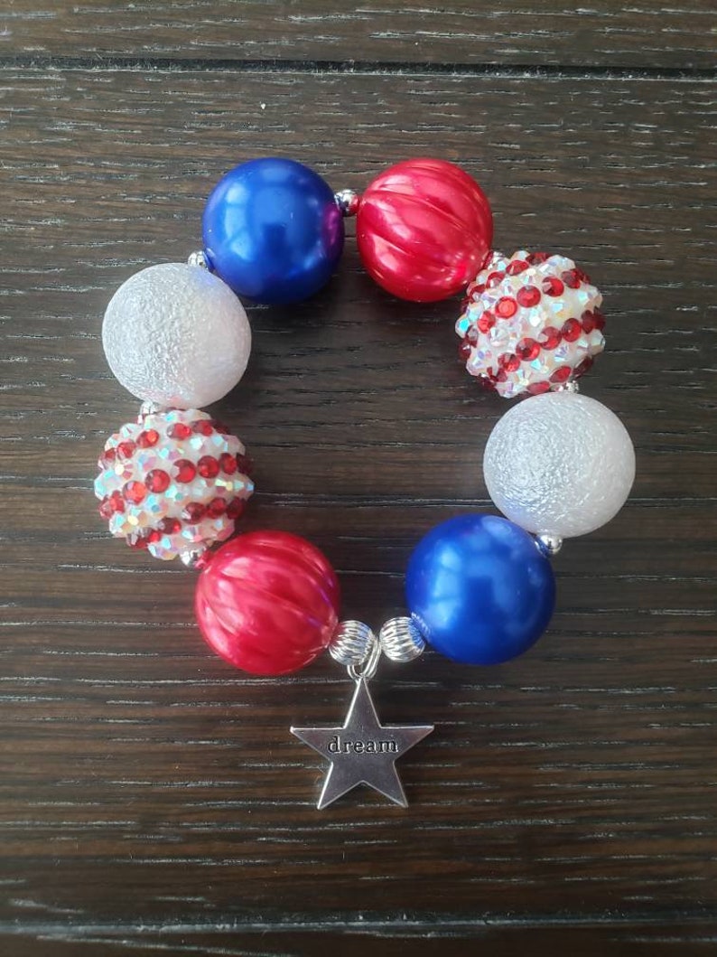May include: A bracelet with red, white, and blue beads, some plain and some decorated with rhinestones. A silver star charm with the word "dream" is attached. The bracelet's design evokes a patriotic theme.