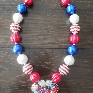May include: A necklace with red, white, and blue beads, featuring a Mickey Mouse pendant. The beads are round and smooth, with some having a striped pattern. The pendant is adorned with red, white, and blue rhinestones. The necklace is secured with a silver clasp.
