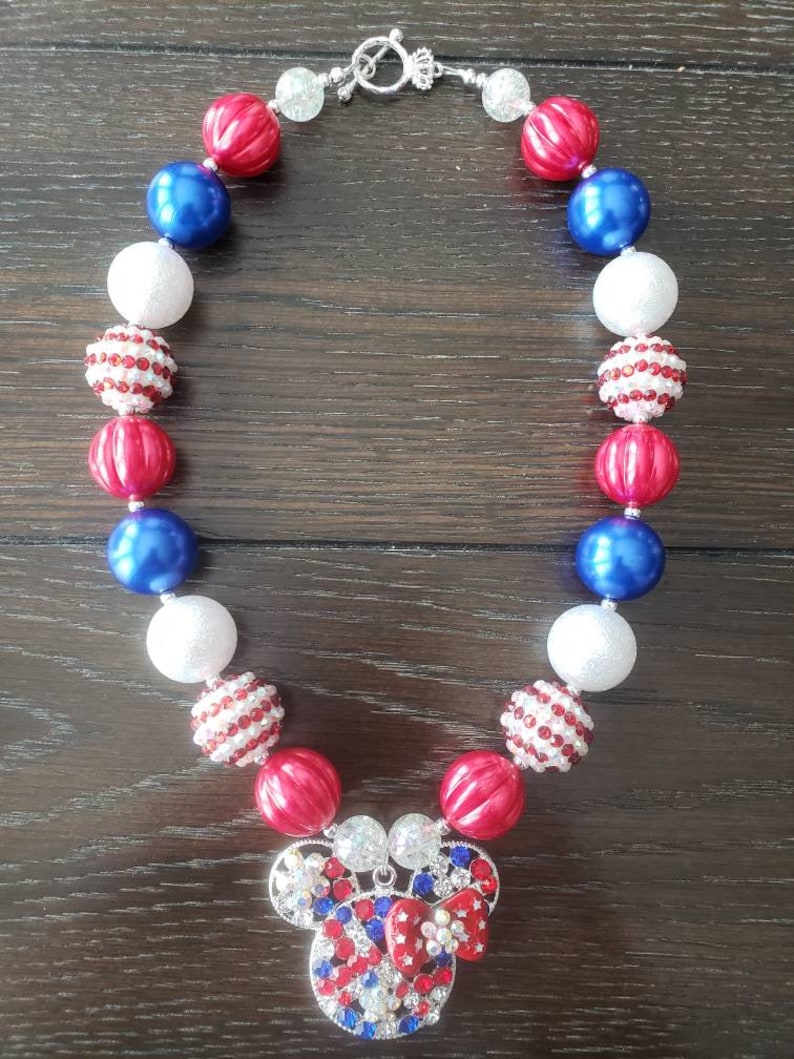 May include: A necklace with red, white, and blue beads, featuring a Mickey Mouse pendant. The beads are round and smooth, with some having a striped pattern. The pendant is adorned with red, white, and blue rhinestones. The necklace is secured with a silver clasp.
