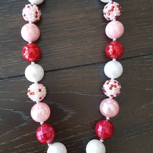 Pode incluir: Um colar de contas grossas vermelho, branco e rosa com um amuleto de Minnie Mouse. O amuleto &eacute; feito de strass e tem um la&ccedil;o rosa.