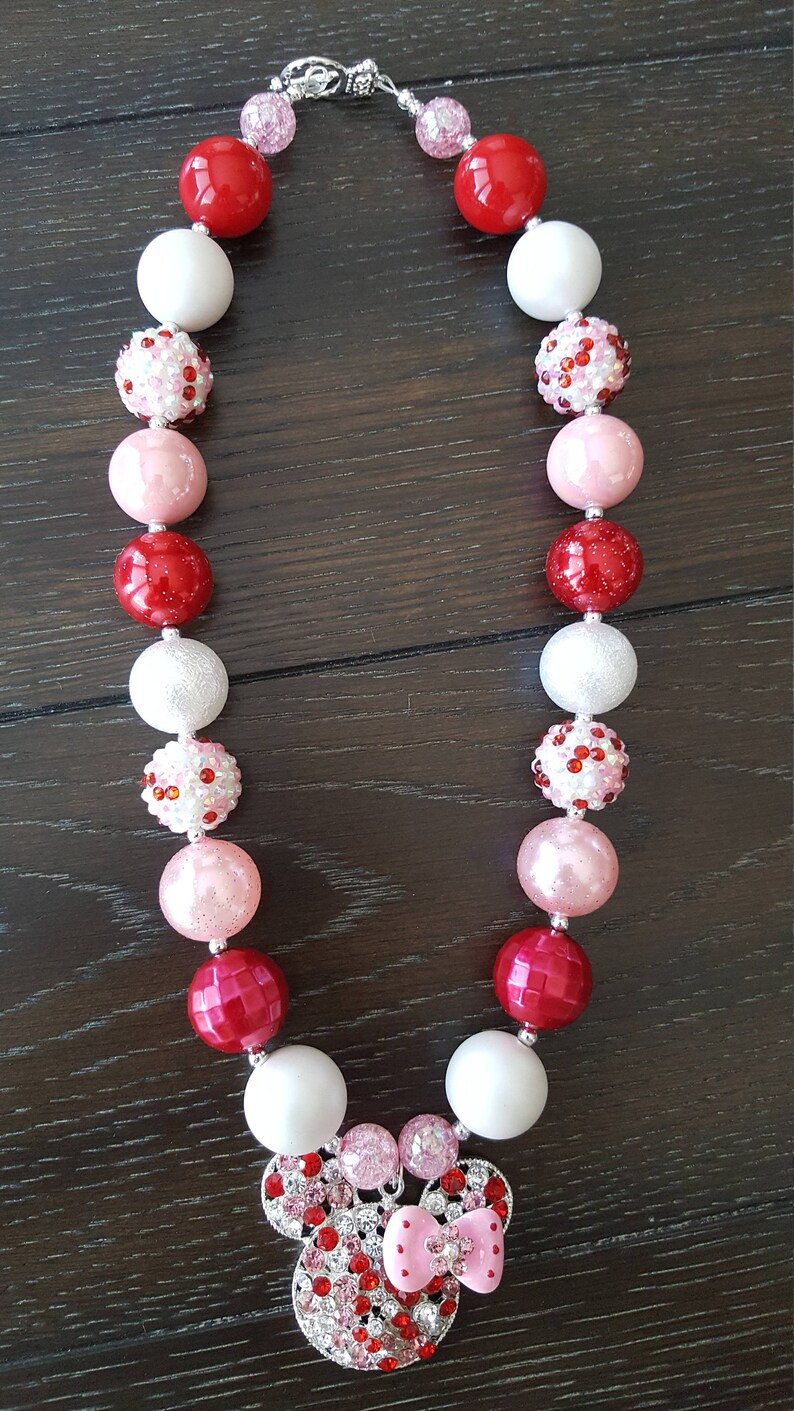 Pode incluir: Um colar de contas grossas vermelho, branco e rosa com um amuleto de Minnie Mouse. O amuleto &eacute; feito de strass e tem um la&ccedil;o rosa.