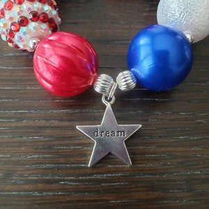 May include: A bracelet in red, white, and blue, with a silver star charm that says "dream". The beads are different sizes and textures. A patriotic accessory. The bracelet is designed to be worn as a fashion item.