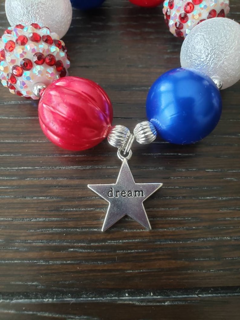 May include: A bracelet in red, white, and blue, with a silver star charm that says "dream". The beads are different sizes and textures. A patriotic accessory. The bracelet is designed to be worn as a fashion item.