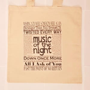 May include: A beige tote bag with a black and white print of lyrics from the musical "The Phantom of the Opera". The lyrics include "Wishing you were somehow here again", "Stranger than you dreamt it", "Twisted every way", "Music of the night", "Down once more", "All I ask of you", and "Past the point of no return".