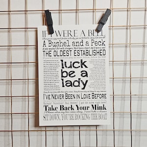 Puede incluir: Una impresión blanca con texto negro, sujeta por dos clips negros sobre una rejilla de alambre dorada. La impresión presenta letras de canciones de "Guys and Dolls", incluyendo "Luck Be a Lady" y "Take Back Your Mink".